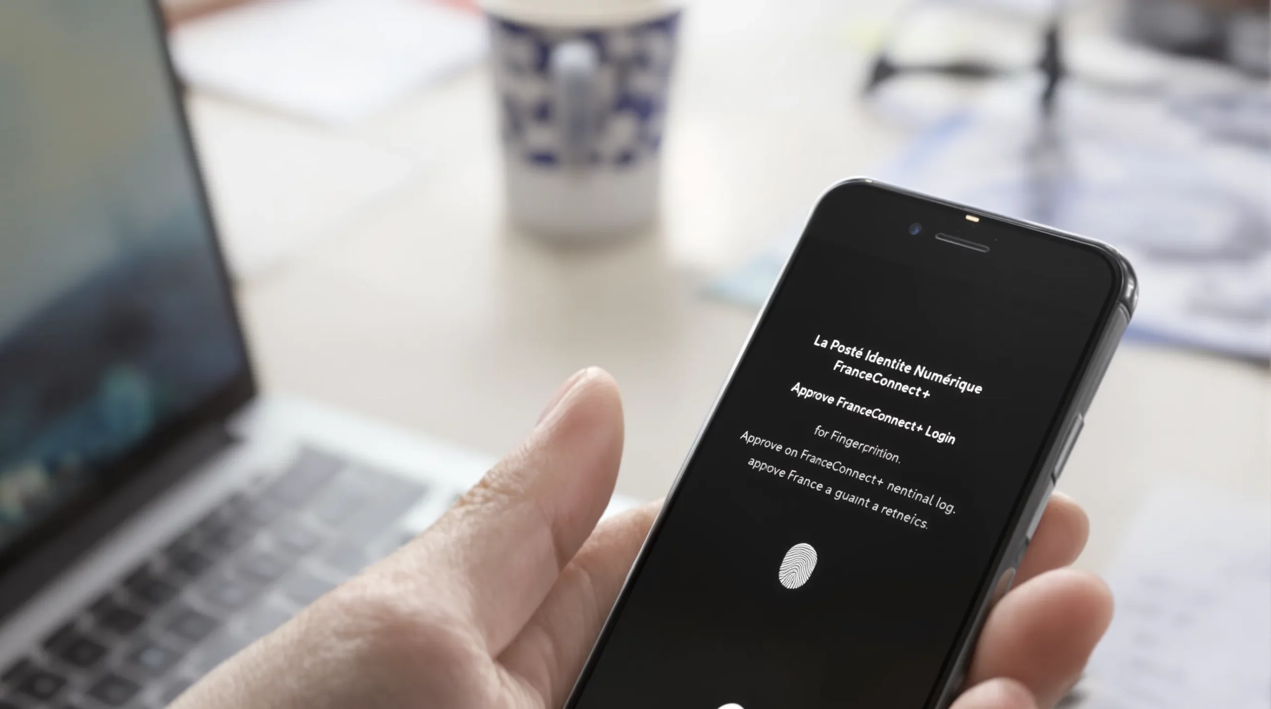 Close-up of a smartphone screen showing the La Poste Identité Numérique app requesting fingerprint authentication to approve a FranceConnect+ login, with a blurred desk in the background.